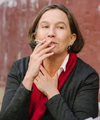 一位留短发的女性近照,她身穿深色外套、系红色围巾,叼着香烟,目光望向一侧。
