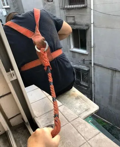 一位维修师傅背对镜头坐在居民楼窗外空调外机旁的窄台上,身系橙色安全绳,画面下方一只手正拽住垂下的安全绳