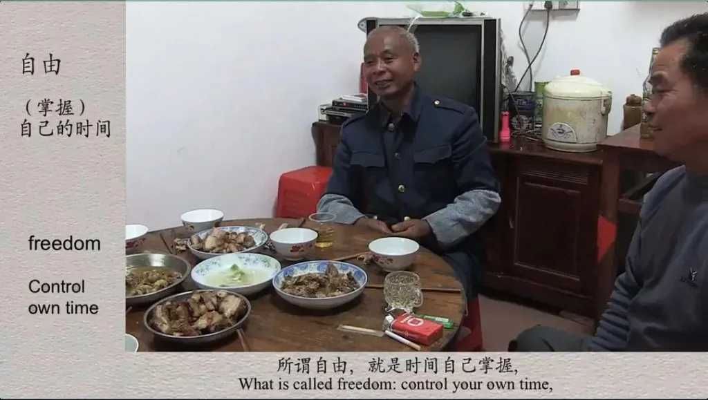视频截图,两位男子坐在摆满家常菜肴的八仙桌旁交谈,画面左侧叠加文字“自由(掌握)自己的时间”及英文翻译,底部字幕写着“所谓自由,就是时间自己掌握”。