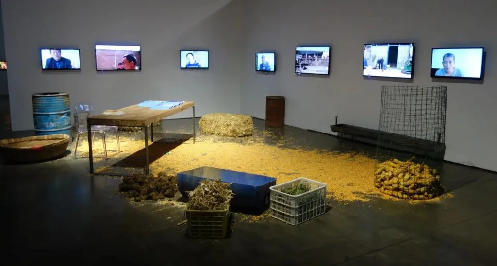 展厅一角,墙上挂着七块显示屏播放访谈影像,地面铺满玉米粒与稻草,散置着塑料筐、木箱、铁桶和一张写字桌,构成装置现场。
