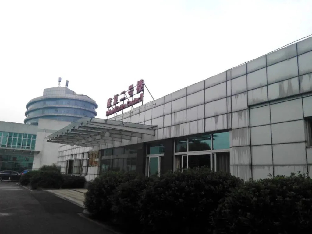 一栋米白色外墙的低层建筑,入口上方挂着牌匾,旁边是一座圆形玻璃塔楼,前方是停车场与灌木丛,天空阴沉。