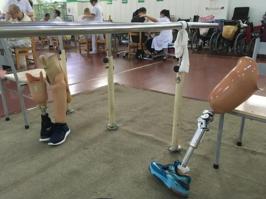 训练室内两具假肢斜靠在金属平行杠上,一具穿深色运动鞋,另一具带外露金属膝关节、穿蓝色运动鞋,背景是坐在桌旁的人们和停放的轮椅。