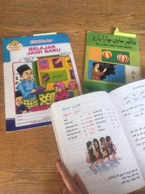 画面中摆着两本封面不同的爪夷文教材,其中一本封面是戴头巾的女性在黑板前书写的插画;下方一只手翻开的课本页面印着爪夷文与罗马字对照词汇表,插图为几名戴头巾的女孩结伴走路上学。