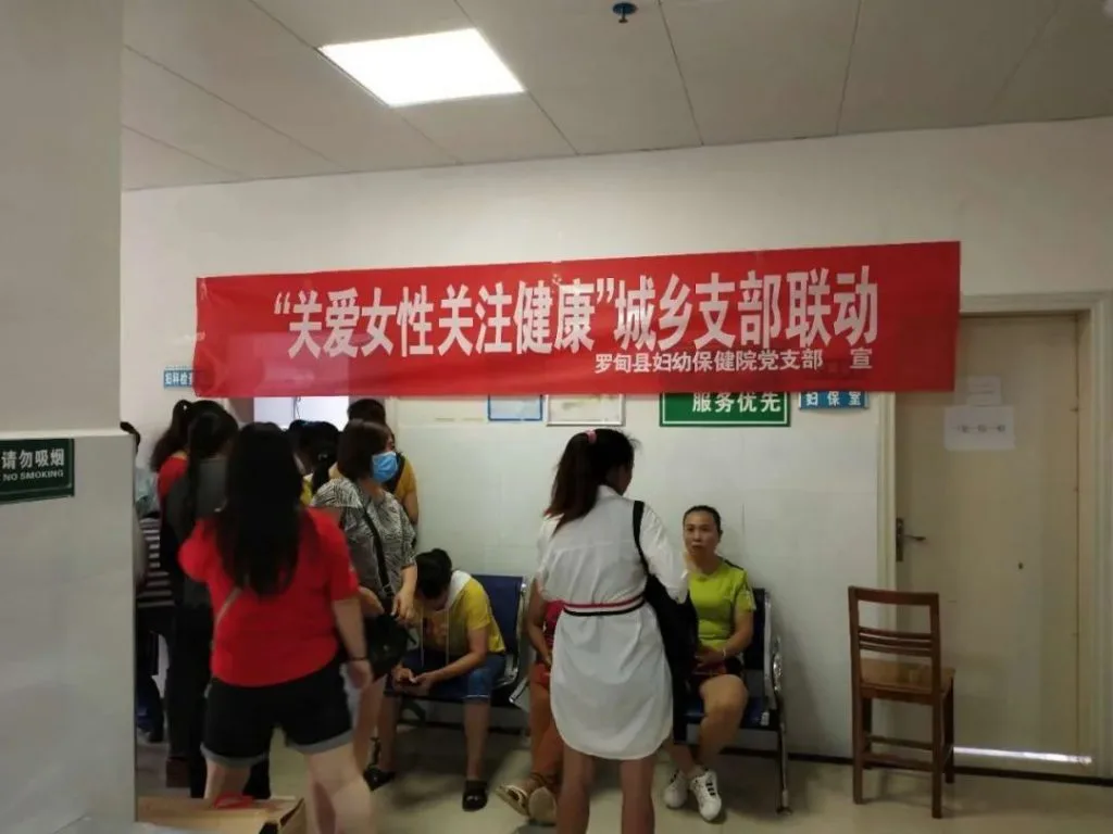 医院走廊里多名女性排队等候,一名穿白大褂的医护人员俯身与一位坐着的女性交谈,头顶悬挂红色横幅,写着“关爱女性关注健康”城乡支部联动。