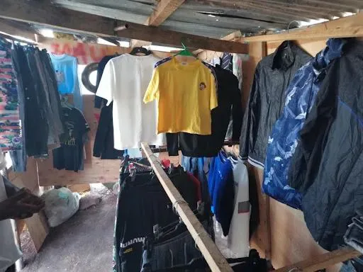 简易木棚搭建的服装摊位内部,横梁和绳索上挂满T恤、外套、牛仔裤等各式衣物,棚顶用木条和铁皮搭成。