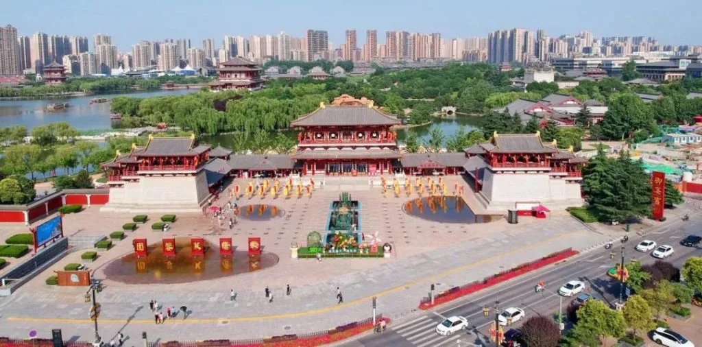 鸟瞰仿唐建筑群与广场,表演者在广场上列队表演,背景是城市高楼、湖泊与绿地。