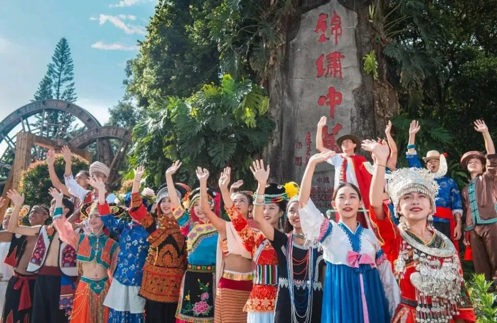 一排身着不同少数民族服饰的表演者面向镜头挥手微笑,背后是刻着“锦绣中华”字样的巨石与水车造型景观。