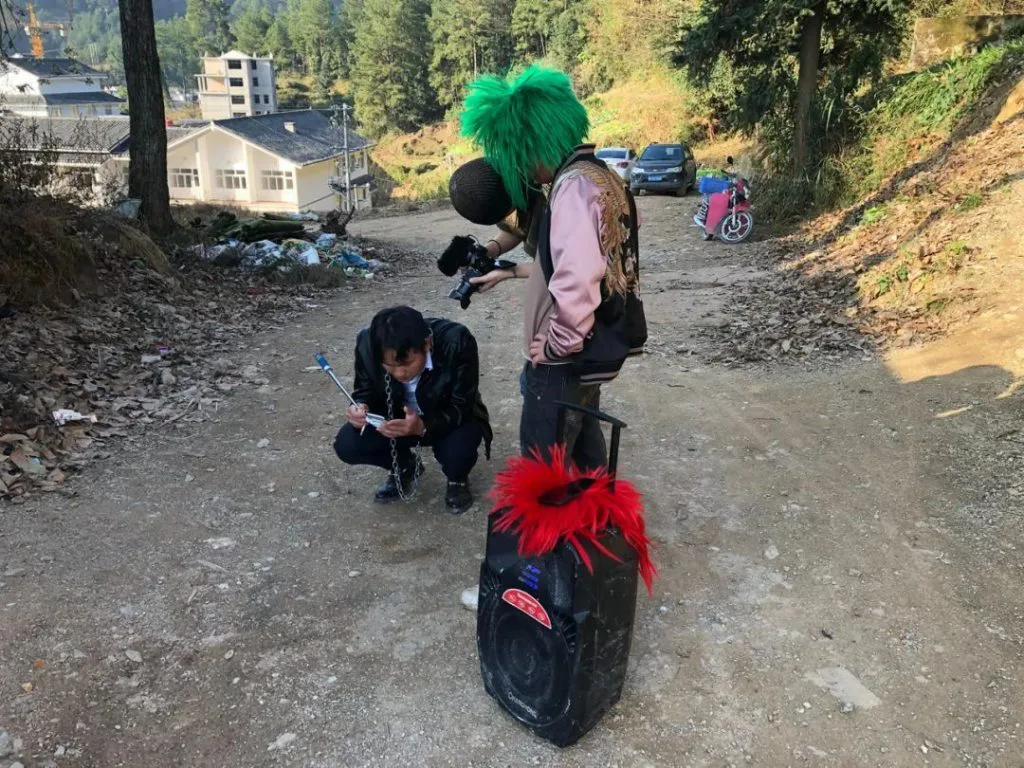 一位头顶绿色蓬松假发、面部戴黑色圆罩的男子手持云台拍摄,脚边放着一个装饰红色羽毛的音箱,另一人蹲地调整设备,背景是山村土路与民房。