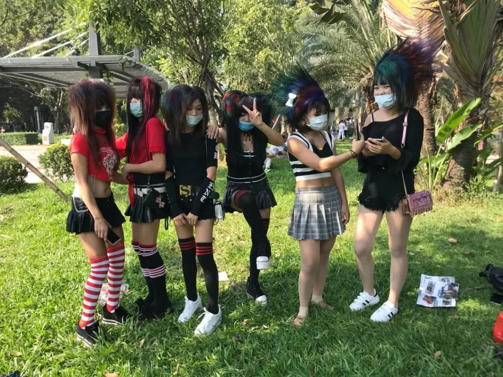 六位染发梳理成蓬松夸张造型的年轻女性戴着口罩,穿着以黑色为主的短裙短裤站在草坪上合影,其中一人比出剪刀手。