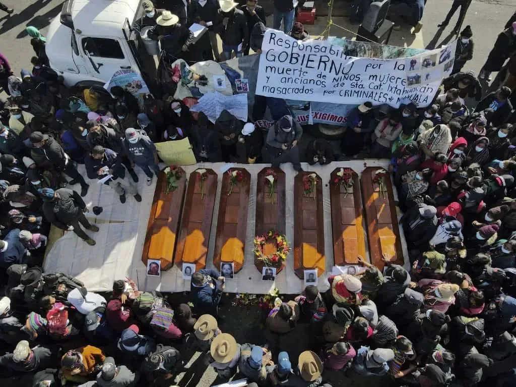 俯瞰视角,七具棺材整齐排列在地面,棺盖上放着遇难者照片和花束,周围站满悼念民众,背景高举一幅西班牙语横幅,谴责政府对儿童、妇女和老人遭黑帮屠杀负有责任。