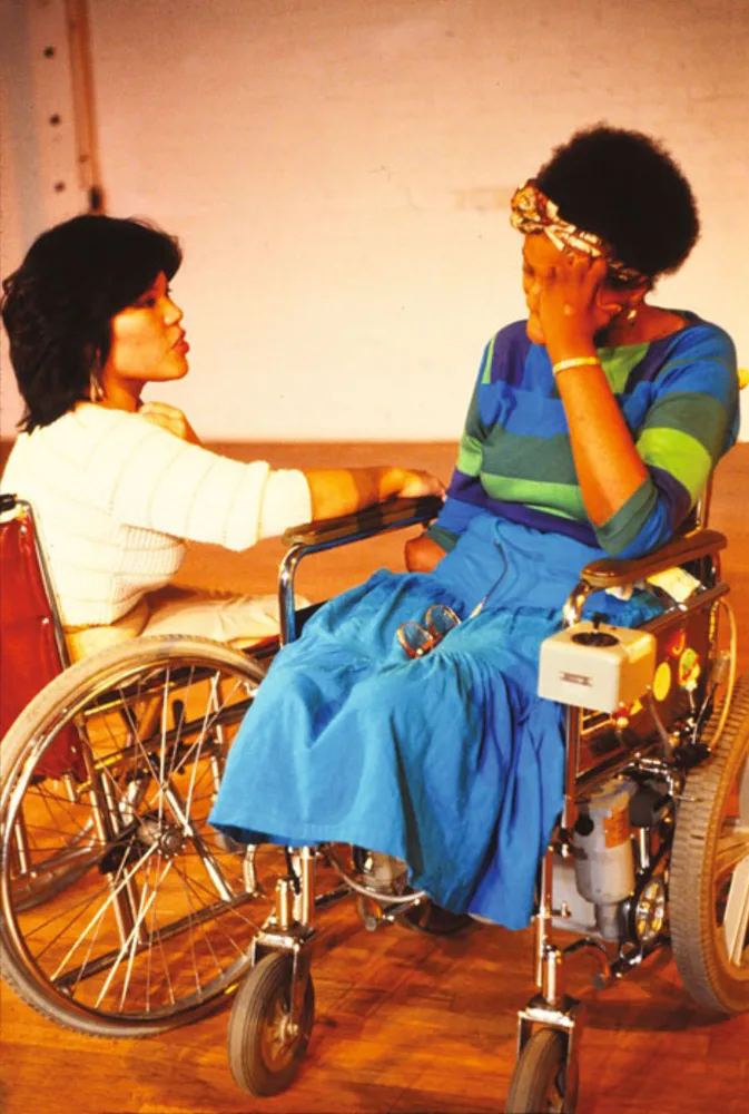 两位坐轮椅的女性在工作室内相对而坐,一人穿白色上衣伸手轻触对方手臂,另一人穿蓝绿条纹裙装,手扶额头低头沉思。