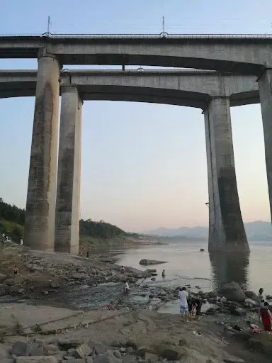 从另一角度拍摄的铁路桥墩,桥下河滩上多人戏水休憩,江面开阔,远处群山连绵。