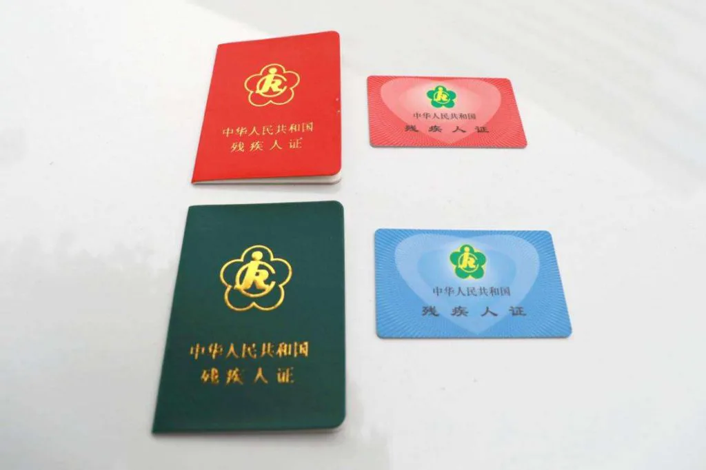 两种版式的残疾人证并排摆放:红色和深绿色的证件本,以及粉色和蓝色的卡片式证件,均印有同一朵小红花徽标。
