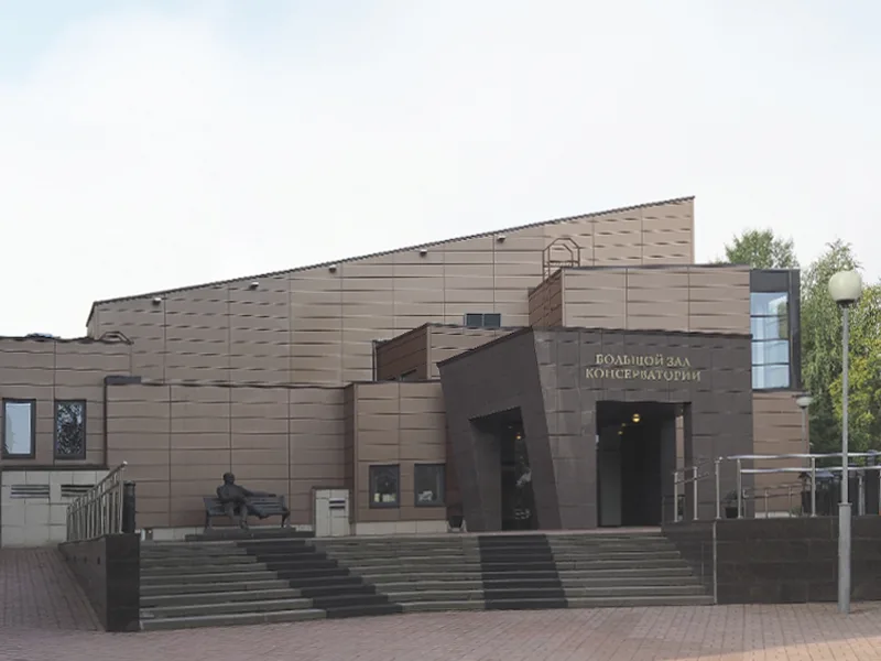 深色几何造型的音乐厅建筑外观,正门前有宽阶梯,一侧另设带扶手的坡道,台阶旁有一尊坐姿人像雕塑。