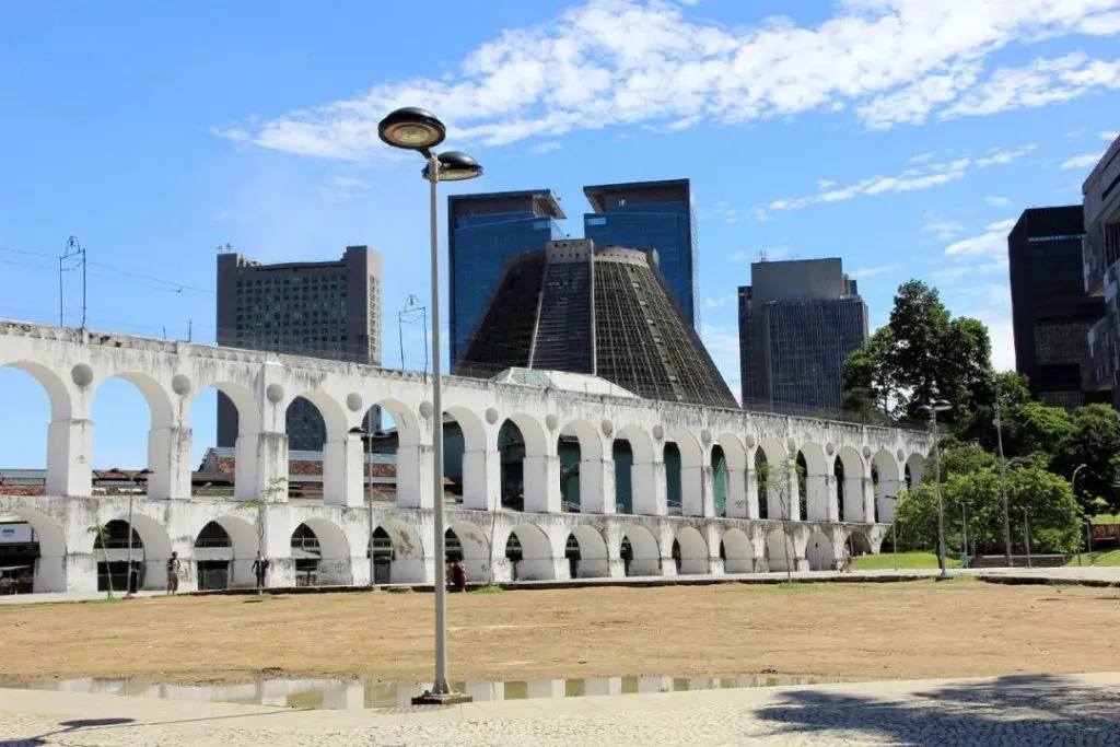白色双层连拱建筑绵延于开阔广场旁,背景是里约现代高楼和圆锥形的都主教座堂,前景地面有积水倒影。