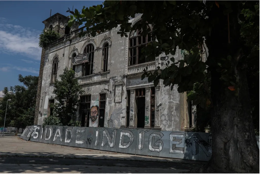 一栋门窗残破、墙面斑驳的老建筑,外墙涂鸦,墙根立着写有'…SIDADE INDÍGENA'(原住民大学)字样的招牌,墙上还有一幅人像涂鸦。