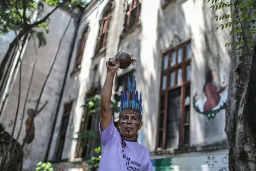 一位脸绘图腾、头戴蓝色羽毛头饰的男性举起手中的沙锤,身后老建筑墙上绘有原住民人物壁画。