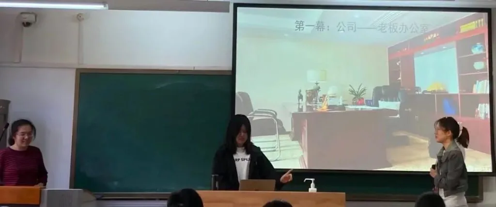 教室里一名学生站在投影屏前讲解,另一名学生站在旁边,老师站在讲台一侧,大屏幕显示'第一幕:公司——老板办公室'字样及办公室场景图,台下学生就座聆听。