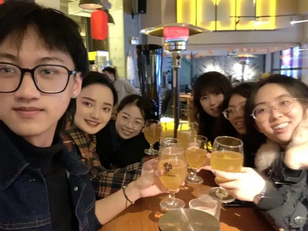 一群年轻人在餐厅围坐一桌,举杯碰杯合影留念,气氛轻松欢快。