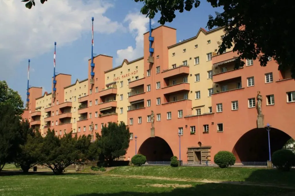 橙黄色相间的狭长住宅楼立面,多座蓝顶塔楼耸立其间,插着旗杆;楼体底层设拱门,前方是草坪与树木。