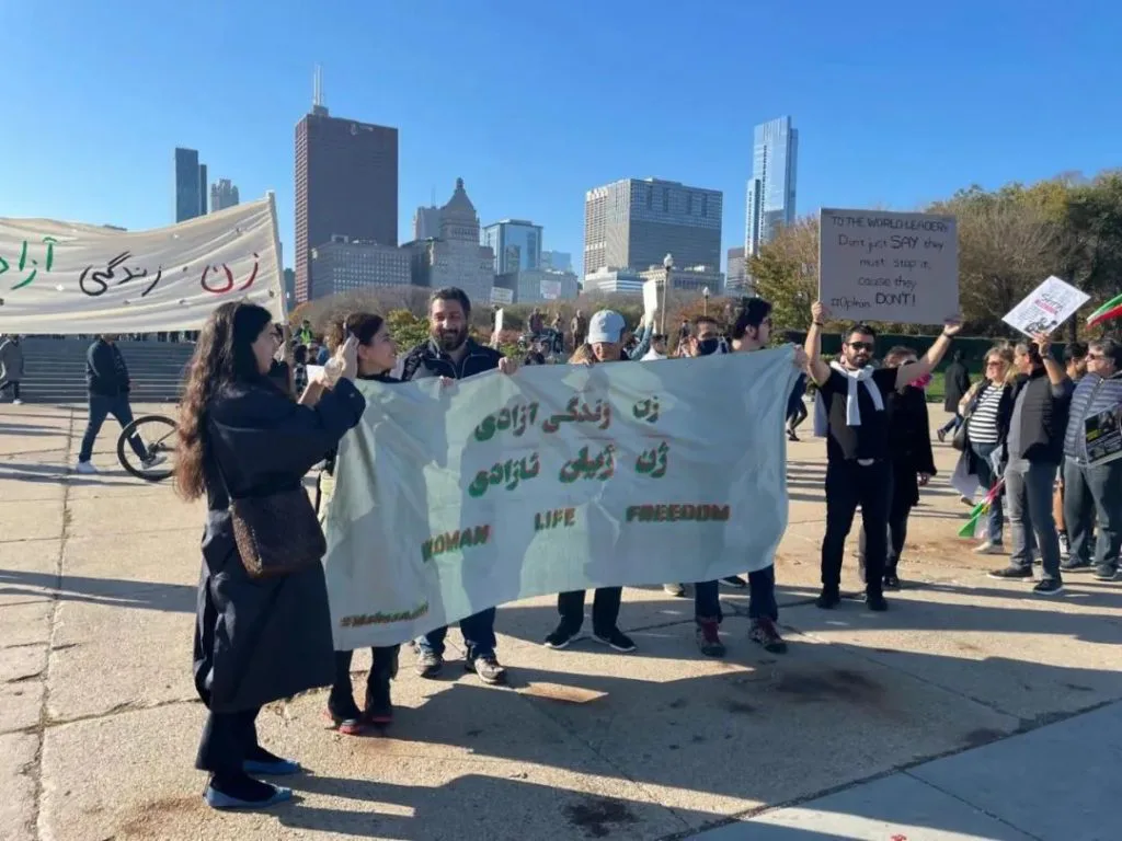 白天户外集会,众人手举写有波斯语和英文“WOMAN LIFE FREEDOM”字样的白色横幅,身后是城市高楼天际线,一旁一人举牌呼吁世界领袖行动。