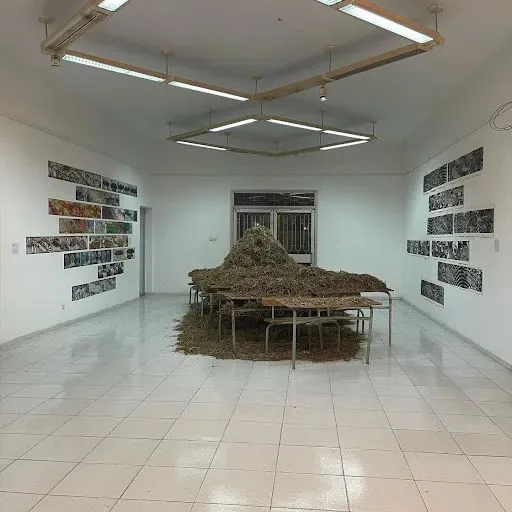 展厅中央多张课桌椅上堆满干草,两侧墙上挂着成排的黑白版画,天花板悬挂着六边形灯具组。