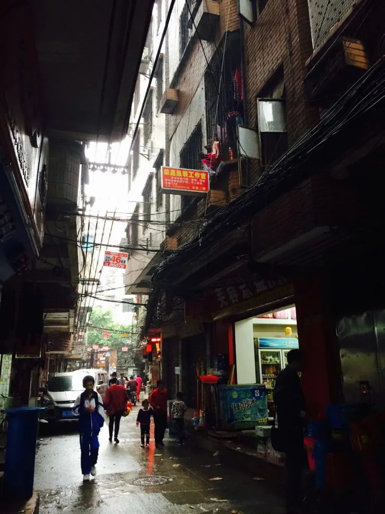 狭窄的城中村巷道两侧高楼相邻紧贴,头顶电线密布,行人和小店铺分布在潮湿的路面两侧
