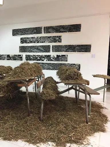 近景可见课桌椅上堆积着大量干草,背景墙上挂着数幅黑白纹理的长条状版画作品。