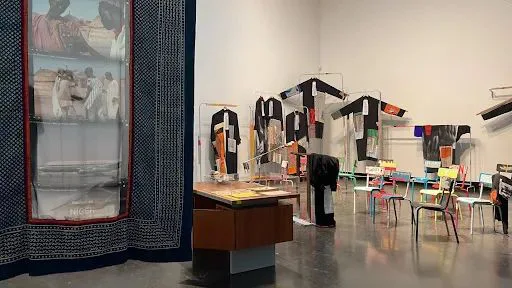 展厅一角悬挂着数件印满图案的黑白拼接长袍,右侧摆放着彩色木椅围绕的桌子,左侧挂帘印有非洲生活场景照片。