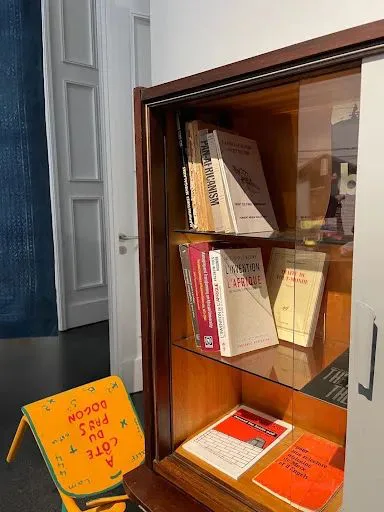 木质玻璃展柜内分层摆放多本书籍,其中可见《L'invention de l'Afrique》,柜前地上放着一把黄色折叠椅,椅面印有法文字样「A côté du pays Dogon」。
