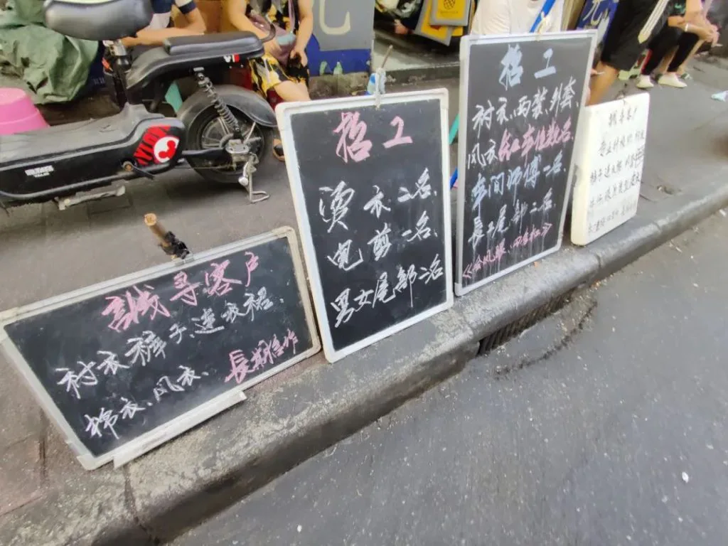 路边并排摆放着四块手写招工黑板,旁边停着一辆电动车,黑板上用彩色粉笔写着不同工种的招工信息,部分字迹潦草难以完全辨认。