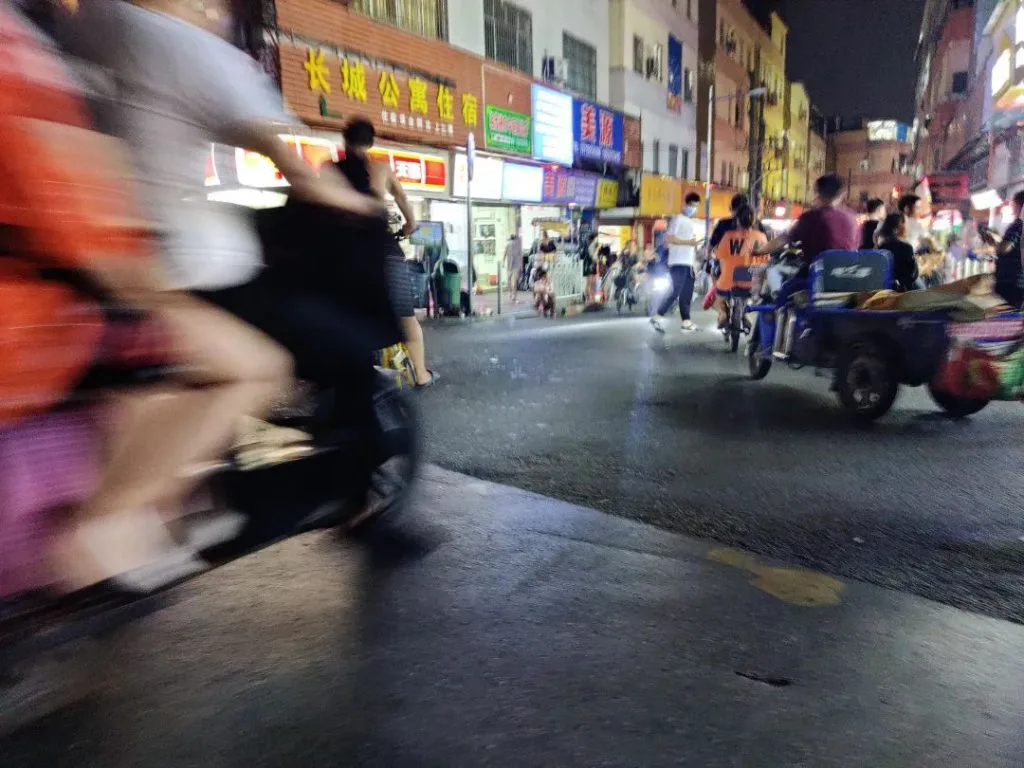 夜间街道两侧商铺灯牌林立,骑手驾驶摩托车快速穿行造成画面模糊,右侧一辆载货三轮车停靠路边,行人往来。