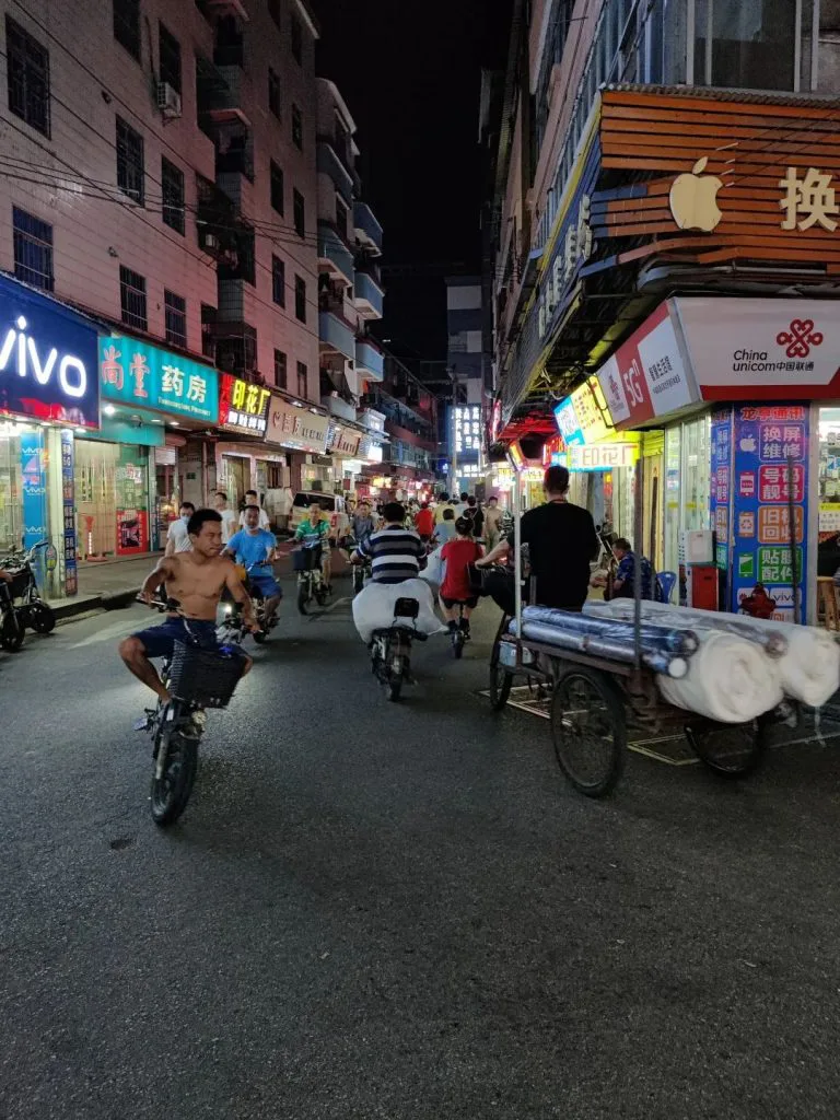狭窄街道两旁手机店、药房等店铺灯光明亮,多名骑手驾驶摩托车穿行,右侧一辆三轮车上堆放着成捆的布匹。