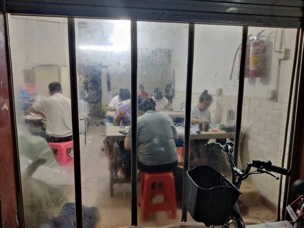 透过卷帘门可见几名工人坐在红色塑料凳上,在长桌前埋头做工,墙上挂着灭火器,门前停着一辆自行车。