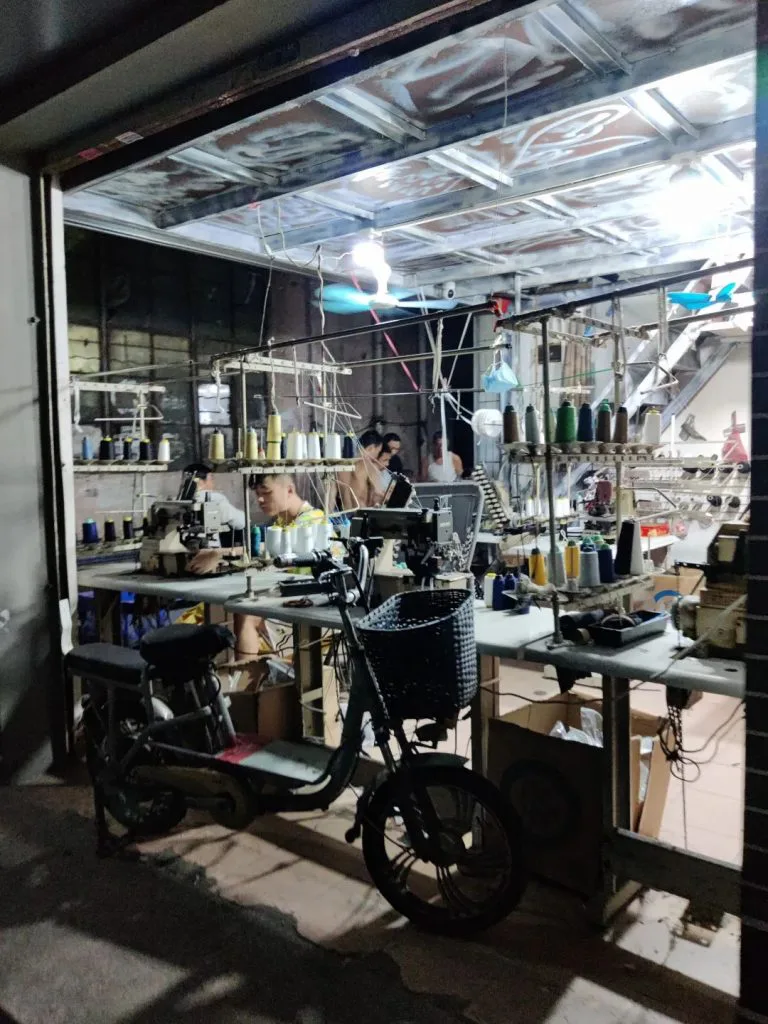卷帘门半开的一楼作坊内,工人坐在缝纫机前工作,身后架子上挂满彩色线轴,门口停着一辆电动车。