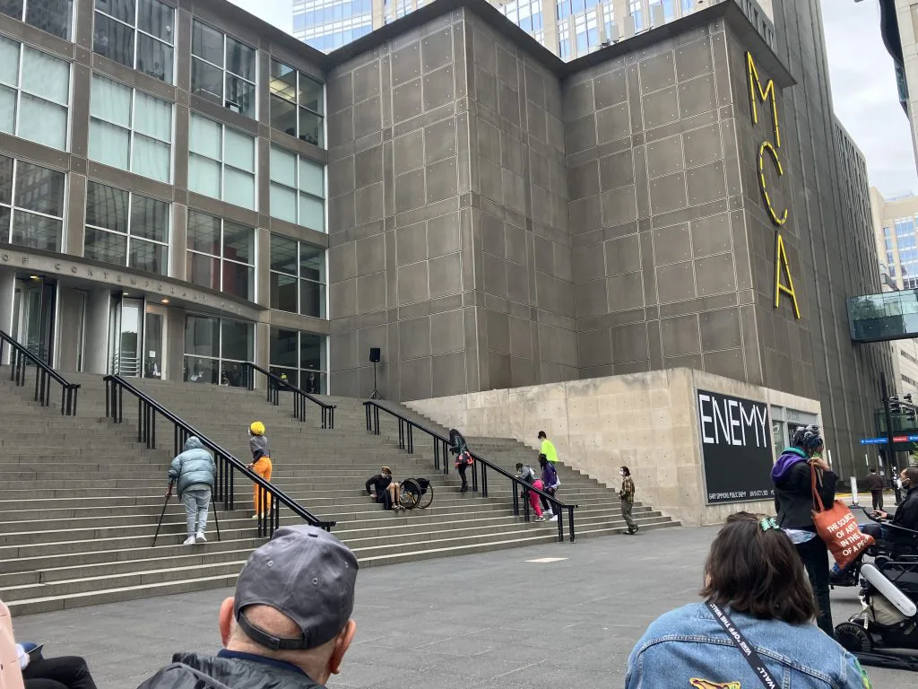 远景,MCA建筑外墙立着黄色巨型'MCA'招牌,宽阔的室外台阶上散布着穿彩色服装的表演者,前景是围观观众的背影。