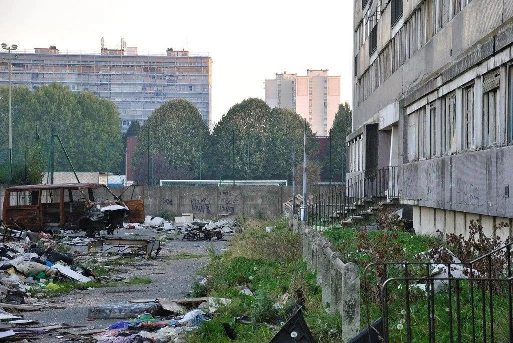 老旧的高层住宅楼下,空地上散落着大量垃圾和一辆烧毁的报废汽车,围墙上布满涂鸦。