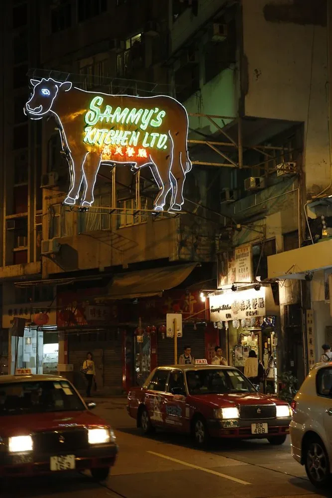 夜晚街道上方悬挂着一块巨大的牛形霓虹招牌,牛身内写着“Sammy's Kitchen Ltd.”及中文店名,下方街道有多辆红色的士驶过。