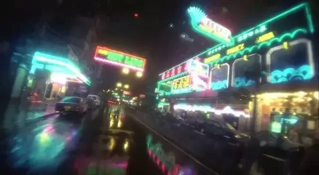 雨夜街景,两侧林立着各色霓虹招牌,湿滑地面倒映着斑斓灯光,一辆的士驶过。