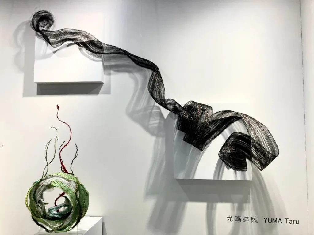 一条黑白编织长带如布匹般从墙上白色展台垂挂盘绕,下方地面摆放一件绿色环状编织物,中心探出几缕暗红色线头,墙面标注艺术家姓名尤玛·达陆。