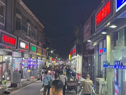 夜晚的皮村商业街灯火通明,两侧店铺霓虹招牌林立,狭窄街道上人潮涌动。