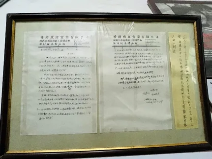 秦梅手写家书原件装裱展示,信纸抬头印着“香港致丽实业有限公司”字样,右侧贴有一张竖写的附言,具体笔迹难以辨认。