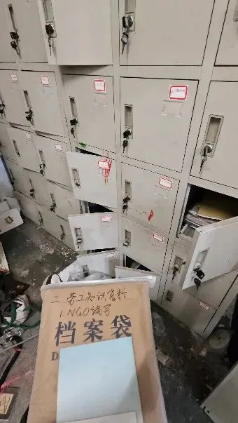 一排铁皮档案柜,多个抽屉贴着红色标签、部分半开可见文件,柜前地上放着写有“档案袋”字样的纸箱,地面散落杂物。