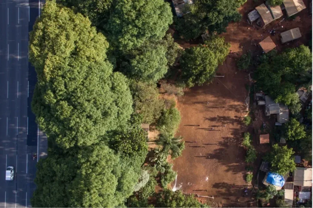 俯瞰图:画面左侧是笔直的多车道公路,右侧红土地上零星分布着简易房屋,被茂密树林环绕,公路与聚居地紧邻。