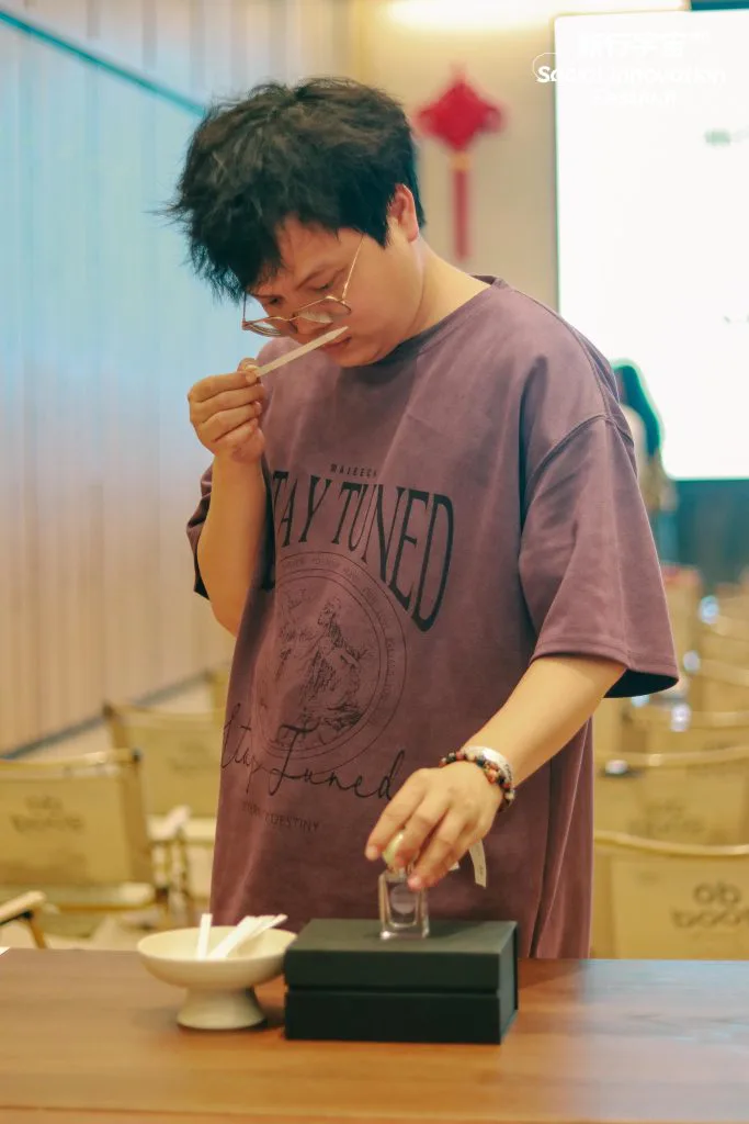 一位戴眼镜、穿紫色印字T恤的人俯身闻嗅试香纸,另一只手拿着小香水瓶,桌上放着白色试纸碗和黑色展台盒。