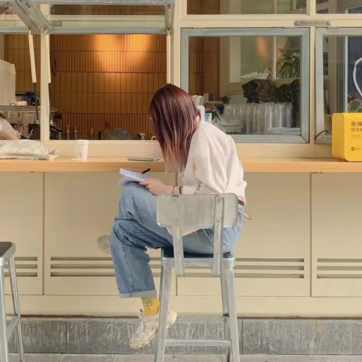 一位长发女性背对镜头,坐在高脚椅上手持纸笔书写,面前是米色吧台和玻璃橱窗,橱窗内可见咖啡机与绿植。
