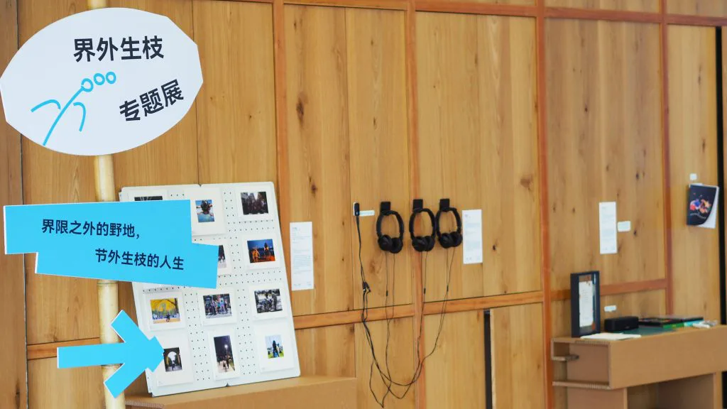 展览现场一角,木质墙面上挂着“界外生枝专题展”圆形指示牌和三副黑色耳机,旁边白色展板陈列着现场照片,墙角桌上摆放着展品。