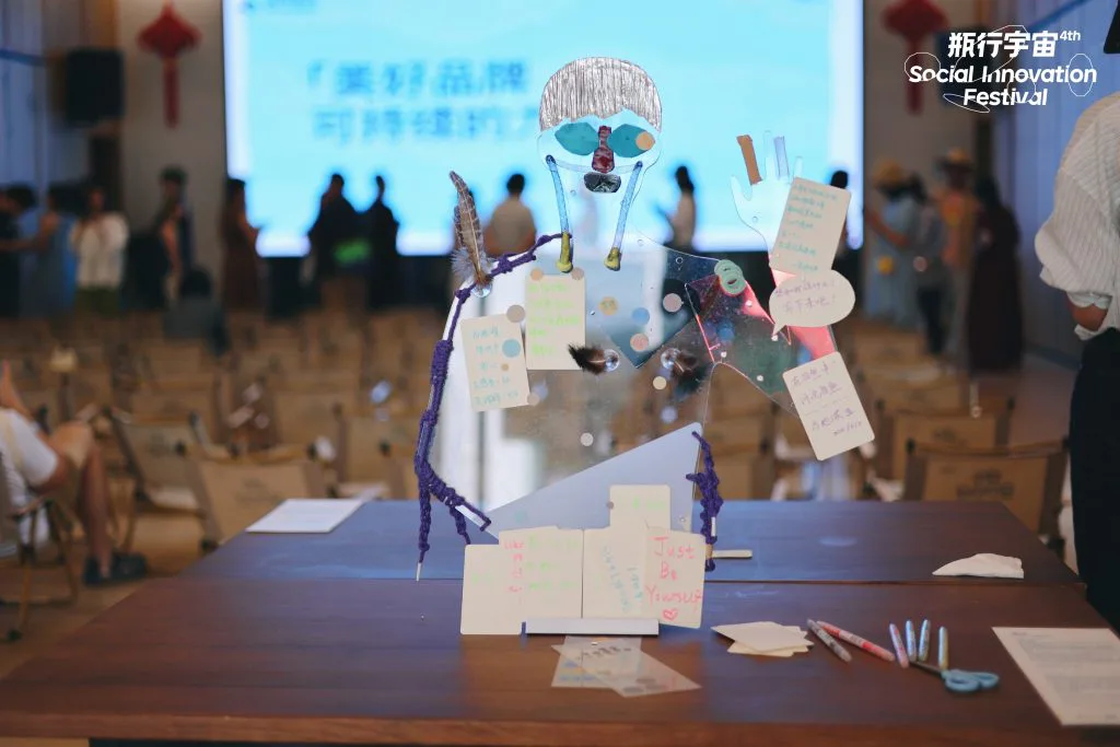 桌上立着人形透明亚克力装置,绘有五官并伸出双臂,身上系着绳索、锡箔、卡纸等材料并挂着多张手写留言卡;背景是展览会场,蓝色大屏幕前坐着观众。