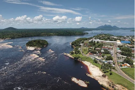 俯瞰视角下,宽阔的黑河中散布着礁石激流,河岸一侧是茂密雨林,另一侧是沿河而建的城镇房屋,远处可见山峦。