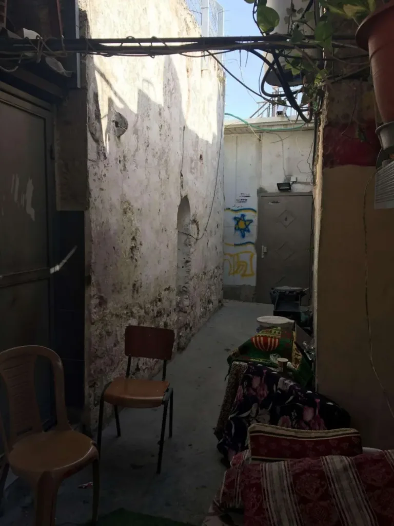狭窄的巷道两侧是斑驳墙壁,一把木椅摆在空地上,尽头一扇门上贴着蓝色大卫星贴纸,右侧堆放着彩色坐垫和布料。